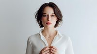 Young woman with short brown hair wearing white shirt against neutral background, looking confidently at camera with natural makeup.