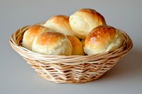 Fresh homemade dinner rolls with golden brown crust served in traditional woven wicker bread basket against neutral background, close up view of baked goods.