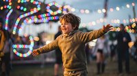 Happy young boy with arms outstretched enjoying festive lights at evening carnival or fair celebration with colorful bokeh background creating joyful atmosphere.