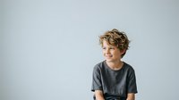 Young boy with light brown wavy hair and fair complexion wearing gray t-shirt, smiling while looking to the side against white background. Suitable for child portraits, family content, and educational materials.