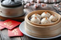 Traditional steamed dumplings in bamboo basket with red envelopes and cherry blossom branches on wooden table, celebrating lunar new year festivities.