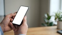 Male hand holding smartphone with blank white screen in modern office workspace with potted plant and wooden desk.