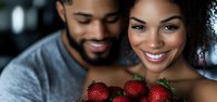 Young African American couple sharing fresh strawberries with joyful expressions. Close-up portrait showing genuine happiness and romantic connection in natural lighting.
