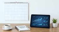 Modern workspace with digital tablet displaying financial analytics dashboard, wall calendar, coffee cup and small plant on wooden desk for business planning.
