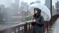 Young woman with dark hair and olive complexion, wearing black rain jacket, holding transparent umbrella on urban bridge during rainy weather with city skyline background.