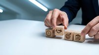 Caucasian businessman arranging wooden cubes with business icons representing time management, partnership, and goal planning on white desk.