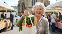 Senior Caucasian woman with gray hair smiling while holding reusable shopping bag with fresh vegetables at outdoor farmers market with church background.