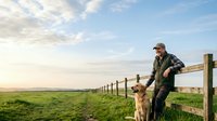 Middle-aged male farmer in work clothes and cap standing by wooden fence with golden retriever dog in rural pasture landscape during golden hour, suitable for agriculture and lifestyle content.