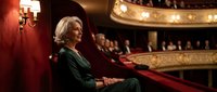 Elegant senior woman in green dress sitting in ornate theater balcony with red velvet seats and golden decorative details during evening performance.