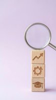 Magnifying glass examining wooden blocks with growth arrow, gear and graduation cap symbols on purple background for business analysis and education concepts.