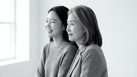 Two professional women colleagues standing together smiling and looking forward in bright office space with natural window light for business teamwork.