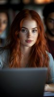 Young redhead woman with freckles working on laptop in modern office environment. Professional female employee focused on computer screen during workday.