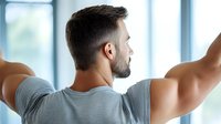 Young Caucasian bearded man in grey t-shirt stretching arms near window in morning, back view. Wellness and healthy lifestyle concept for active people.