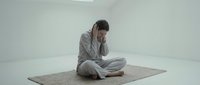 Young woman in gray pajamas sitting cross-legged on woven mat in minimalist white room, holding head in hands showing stress, depression or mental health.