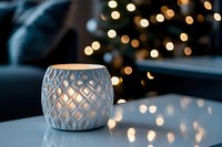 White ceramic candle holder with geometric pattern on table against blurred Christmas lights background creating cozy festive atmosphere.