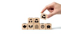 Hand placing wooden cube with coffee cup icon on pyramid of blocks featuring various business symbols on white background for branding concepts.