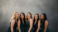Five diverse women in elegant dark green dresses laughing together against smoky gray backdrop for wedding party celebration events.