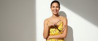 Young woman with dark hair and olive complexion wearing yellow floral dress, smiling at camera against white wall with natural sunlight for fashion, lifestyle, and portrait use.