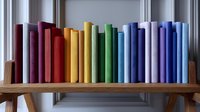 Colorful books arranged in rainbow spectrum order standing on wooden shelf against white wall, creating gradient effect from red through yellow to blue tones.