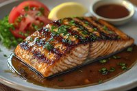 Grilled salmon fillet with teriyaki glaze and fresh herbs, served on plate with sliced tomatoes and lettuce garnish, accompanied by dark sauce.
