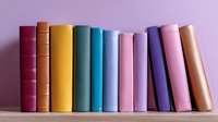 Row of colorful hardcover books standing on wooden shelf against purple wall background, arranged by rainbow spectrum colors for modern home library decor.