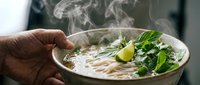 Steaming bowl of noodle soup held in hand with fresh herbs, lime and garnish, ready to serve for Asian cuisine cooking and food preparation.