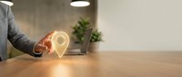 Location pin glowing in hand of businessman at wooden desk with laptop and plant, representing digital navigation, business technology, and global connectivity for corporate applications.