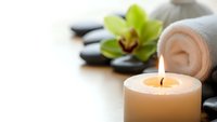 Spa wellness scene with burning candle, rolled towel, hot stones and green orchid flower on wooden surface for relaxation and massage therapy treatments.