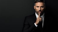 Dramatic portrait of mature Caucasian businessman in black suit making silence gesture against dark background. Professional corporate headshot with moody lighting.