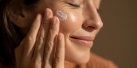 Young woman applying facial cream with fingertips, enjoying skincare routine in warm sunlight. Glowing skin shows hydration benefits.