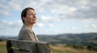 Middle-aged Caucasian woman sitting peacefully on wooden bench overlooking scenic countryside landscape with rolling hills and cloudy sky for wellness content.