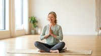 Mature woman with blonde hair, Caucasian ethnicity, wearing glasses and light green cardigan, practicing yoga meditation in lotus position on mat in bright studio space with natural light.