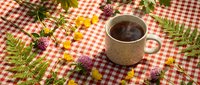 Steaming coffee cup on red checkered tablecloth surrounded by fresh wildflowers and green fern leaves creating cozy morning atmosphere for cafe branding.