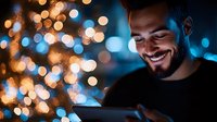 Young bearded man smiling while looking at digital tablet screen at night, illuminated by warm bokeh lights and blue glow creating magical atmosphere.