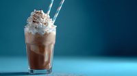 Creamy chocolate milkshake topped with whipped cream and cocoa powder in tall glass with striped straws against blue background, food photography styling.