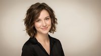 Professional young Caucasian businesswoman with short curly brown hair smiling confidently in black blazer against neutral background for corporate headshots.