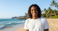 Happy young Black man with dreadlocks smiling on tropical beach with palm trees and ocean waves. Vacation lifestyle portrait for summer travel concepts.