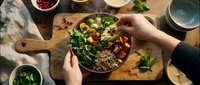 Overhead view of hands holding a wooden bowl with quinoa salad, fresh greens, roasted vegetables and pomegranate seeds on rustic table for healthy eating content.