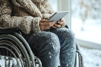 Person in wheelchair wearing cozy beige sweater using digital tablet, view of hands and lap. Snowy winter scene through window creates peaceful atmosphere.