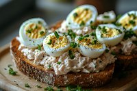 Sliced hard-boiled eggs with tuna salad spread on dark rye bread, garnished with fresh herbs and spices, close-up view of appetizing sandwich.