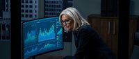 Senior woman with white hair and glasses analyzing financial data on multiple monitors in modern office setting for business analytics and investment research.
