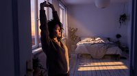 Young woman with dark hair and medium skin tone stretching arms overhead in sunlit bedroom during morning routine, wearing casual striped shirt, surrounded by potted plants and unmade bed.
