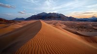 Rippled sand dunes stretch across desert landscape at sunset, dramatic mountain silhouettes on horizon, golden light illuminating textured sand ridges in foreground.