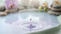 Serene water droplet creating ripples in spa bowl with orchid flowers and stacked stones, wellness and relaxation concept for meditation and spa treatments.