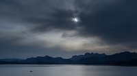 Dramatic stormy sky with dark clouds over mountain range and calm lake water creating moody atmospheric landscape for nature and weather concepts.