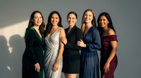 Five diverse young women in elegant evening dresses posing together for group portrait against neutral background in studio lighting.