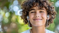 Happy young boy with curly hair listening to music with earphones outdoors, smiling with eyes closed enjoying peaceful moment in natural sunlight.
