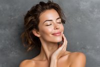 Young woman with glowing skin and closed eyes touching face gently against gray background. Portrays beauty, skincare, and wellness.