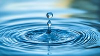 Macro photography of water drop impact creating concentric ripples on blue water surface with second droplet suspended above, capturing moment of collision.