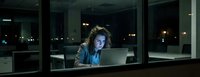 Young professional woman working late at night in modern office with laptop computer illuminated by screen glow against city lights background.